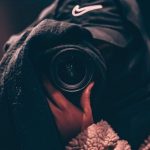 Hidden Business Risks - Unrecognizable person wearing hooded clothes and hiding face behind black scarf taking photo on professional camera on blurred background
