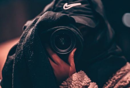 Hidden Business Risks - Unrecognizable person wearing hooded clothes and hiding face behind black scarf taking photo on professional camera on blurred background