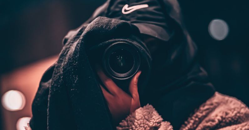 Hidden Business Risks - Unrecognizable person wearing hooded clothes and hiding face behind black scarf taking photo on professional camera on blurred background