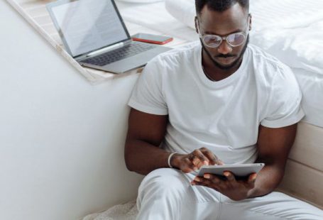 Remote Work - A Man Sitting on the White Blanket on the Floor while Using His Tablet